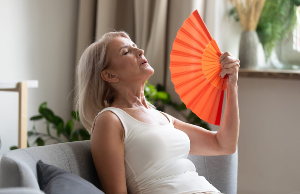 Stressed annoyed old senior woman using waving fan suffer from overheating, summer heat health hormone problem, no air conditioner at home sit on sofa feel exhaustion dehydration heatstroke concept