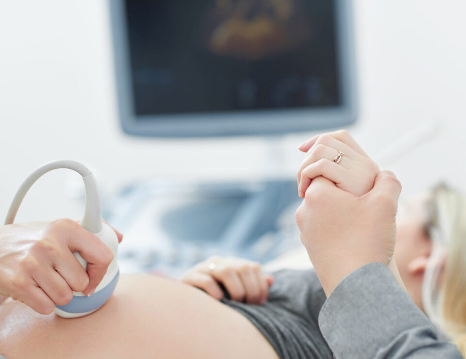 Doctor touching stomach by tool of female who lying and looking at screen of computer. Professional doctor using ultrasound equipment and gel screening of pregnant woman. Husband holding hand of wife.
